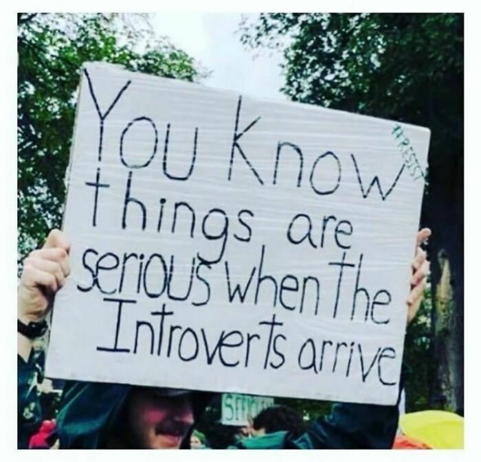 Sign reading "You know things are serious when the introverts arrive," held up at an outdoor event.
