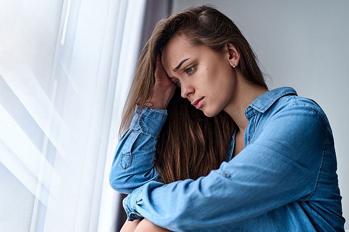 Woman in a blue shirt looking pensive by the window, related to family Thanksgiving invitation dispute. Woman in a blue shirt looking pensive by the window, related to family Thanksgiving invitation dispute.