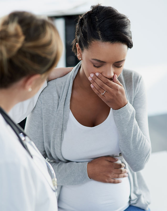 Pregnant woman in a gray cardigan holding her stomach, looking unwell, with a doctor nearby offering support. Pregnant woman in a gray cardigan holding her stomach, looking unwell, with a doctor nearby offering support.