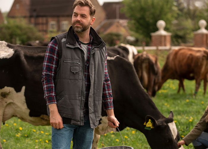 Farmer standing in a field with cows, holding a bucket, smiling confidently in a rural setting. Farmer standing in a field with cows, holding a bucket, smiling confidently in a rural setting.