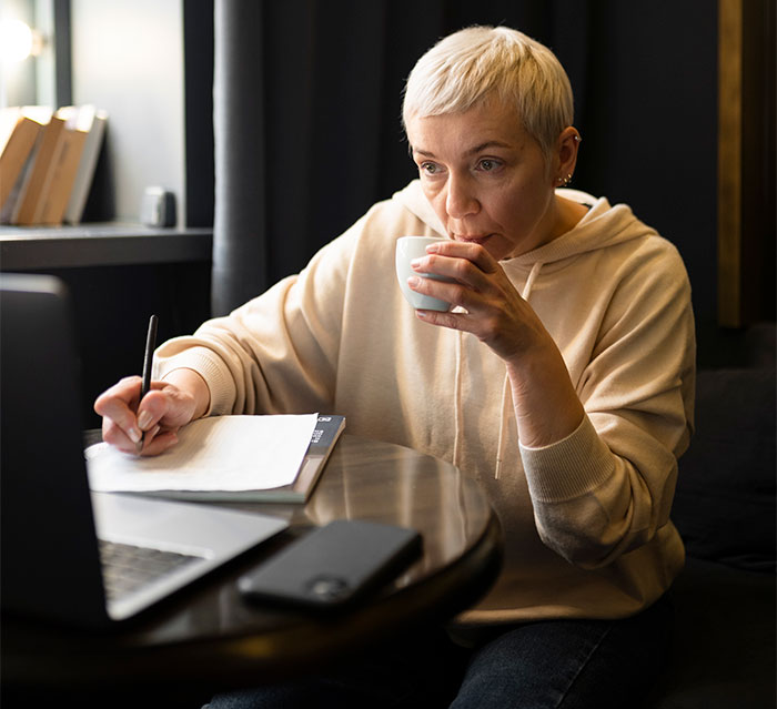 Woman in a beige hoodie sipping coffee, holding a pen, and writing notes near a laptop, conveying suspicion of MIL. Woman in a beige hoodie sipping coffee, holding a pen, and writing notes near a laptop, conveying suspicion of MIL.