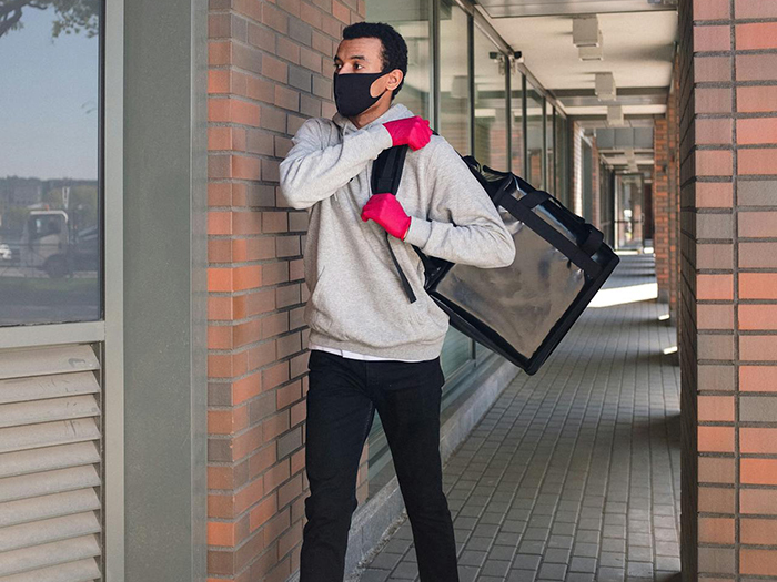 Man wearing mask and pink gloves, carrying a delivery bag near a brick building. Man wearing mask and pink gloves, carrying a delivery bag near a brick building.