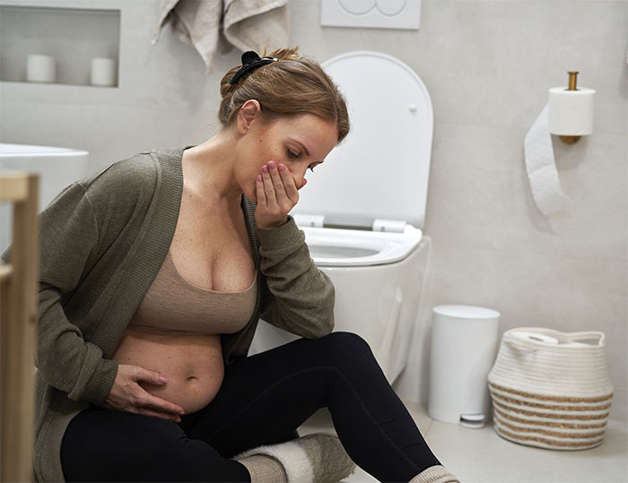 Pregnant woman feeling sick in bathroom, sitting on floor near toilet, holding hand to mouth. Pregnant woman feeling sick in bathroom, sitting on floor near toilet, holding hand to mouth.