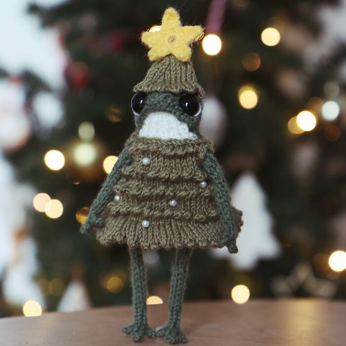 Knitted miniature frog wearing a festive outfit with a star, set in front of a blurred, decorated tree background.