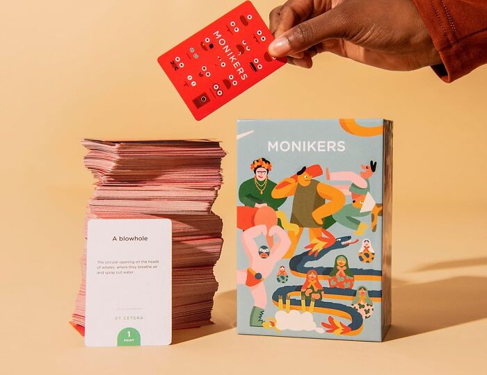 Hand holding a Monikers game card above a stack of cards and a colorful Monikers game box, showcasing compact games.