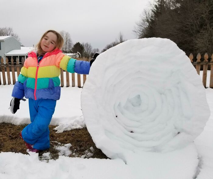 Child in colorful jacket next to large rolled snowball, showcasing ice-cold winter scenery.