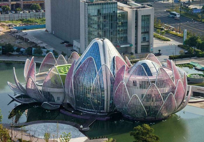 Futuristic flower-shaped building with glass and metal architectural feats reflected in surrounding water near urban area.