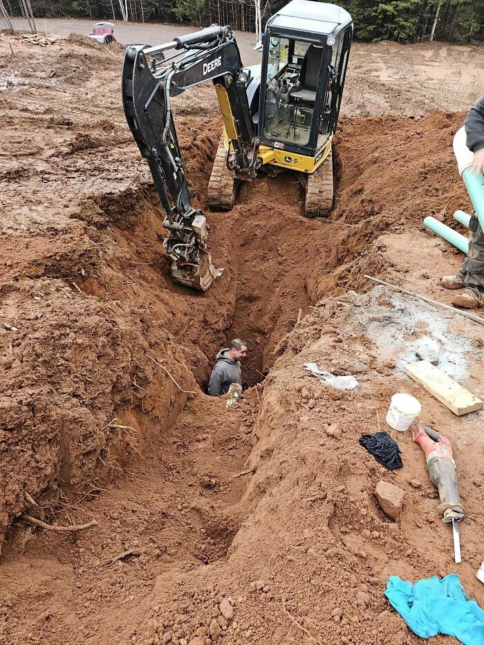 Man standing in deep trench next to excavator, illustrating workers ignoring safety protocol and making dangerous decisions.