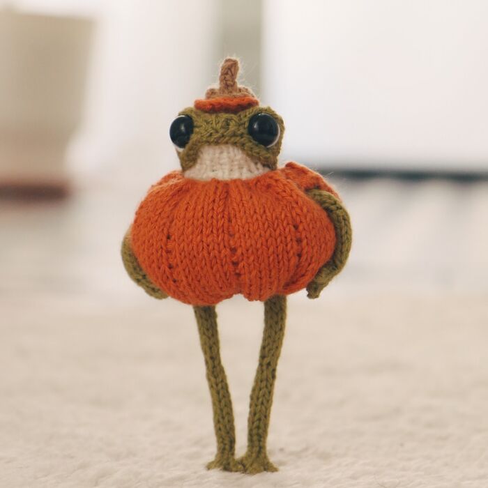 Miniature knitted frog with orange body by a French knitter, standing on a textured surface.