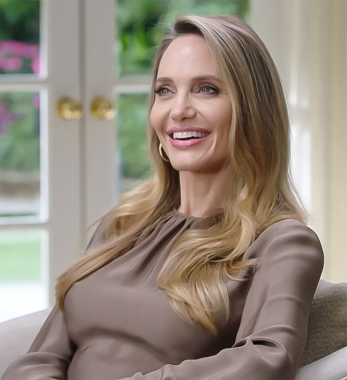 Angelina Jolie with long hair and a warm smile, sitting indoors, dressed in a brown top. Angelina Jolie with long hair and a warm smile, sitting indoors, dressed in a brown top.