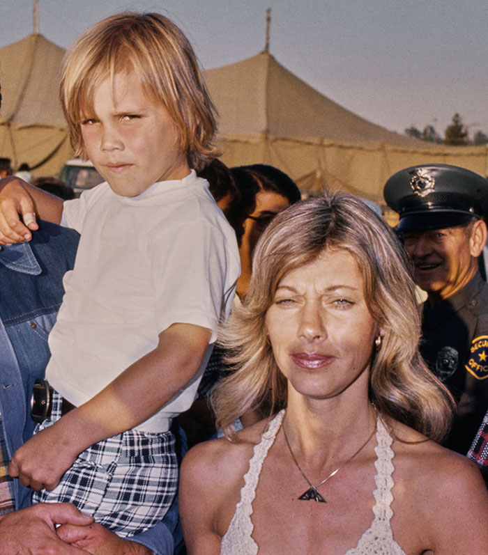 Mother and child outdoors, woman with long blonde hair and boy in white shirt, standing in front of tents and a police officer. Mother and child outdoors, woman with long blonde hair and boy in white shirt, standing in front of tents and a police officer.