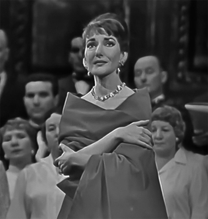 A black-and-white image of Maria Callas in elegant attire, wearing a necklace, standing in front of an audience. A black-and-white image of Maria Callas in elegant attire, wearing a necklace, standing in front of an audience.