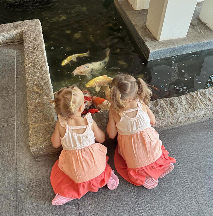 Two young children, connected to Bobbi Althoff’s husband, sitting by a koi pond, wearing matching dresses, gazing at colorful fish. Two young children, connected to Bobbi Althoff’s husband, sitting by a koi pond, wearing matching dresses, gazing at colorful fish.