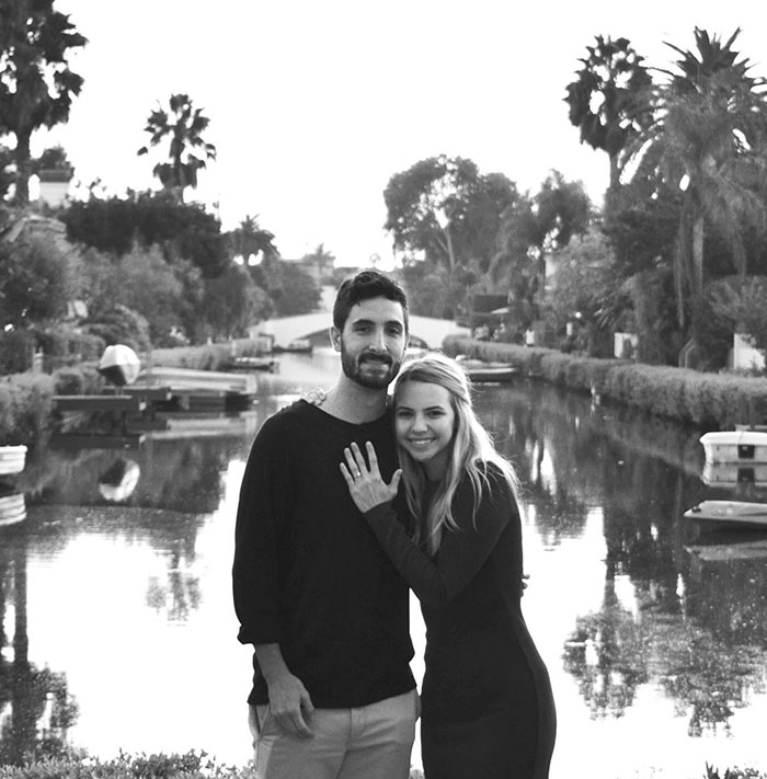 A black-and-white photo of Bobbi and Cory Althoff standing by a serene canal, smiling, surrounded by palm trees and a picturesque bridge. A black-and-white photo of Bobbi and Cory Althoff standing by a serene canal, smiling, surrounded by palm trees and a picturesque bridge.
