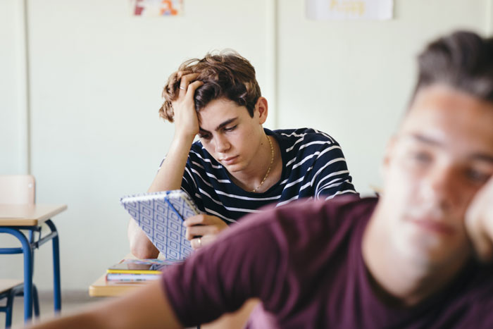 Picked-on teen in classroom silently plotting, notebook in hand, bully unaware in foreground. Picked-on teen in classroom silently plotting, notebook in hand, bully unaware in foreground.