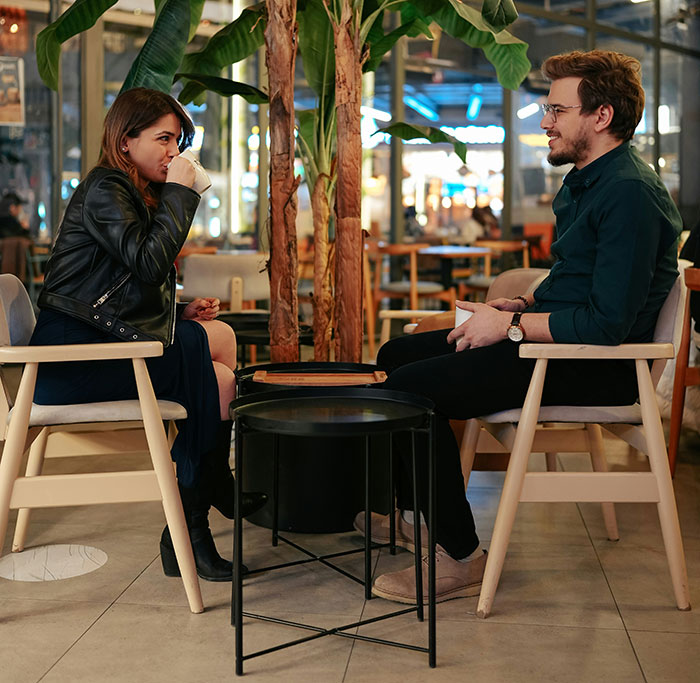 Woman on a date in a cafe, sipping coffee, with a man talking, surrounded by plants in a cozy setting. Woman on a date in a cafe, sipping coffee, with a man talking, surrounded by plants in a cozy setting.