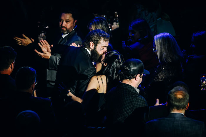 A candid moment of Post Malone embracing his fiancée in a dimly lit event setting, surrounded by a seated crowd in formal attire. A candid moment of Post Malone embracing his fiancée in a dimly lit event setting, surrounded by a seated crowd in formal attire.