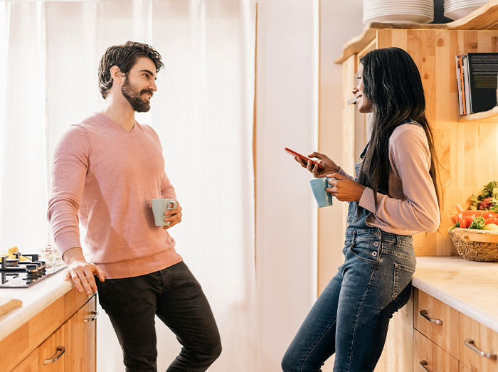 Man and woman chatting in a kitchen, both holding mugs, amidst a bright and cozy setting. Man and woman chatting in a kitchen, both holding mugs, amidst a bright and cozy setting.
