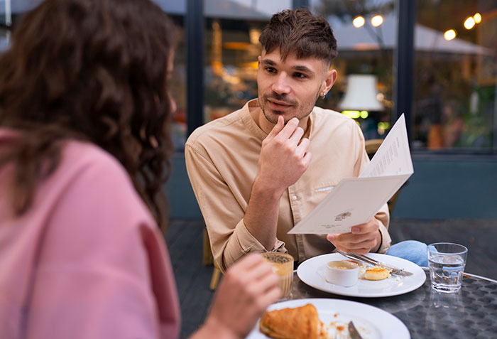 A woman with a meat burger and a man reading the menu at an outdoor restaurant table, highlighting a dietary difference on a date. A woman with a meat burger and a man reading the menu at an outdoor restaurant table, highlighting a dietary difference on a date.
