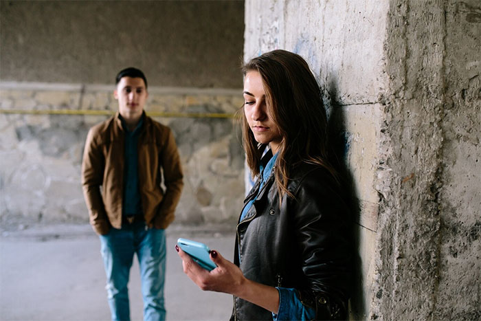 Woman leans against a wall, looking at her phone, while a man stands in the background. Woman leans against a wall, looking at her phone, while a man stands in the background.