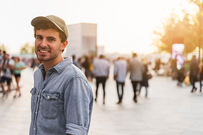 Man in a cap and denim shirt smiling, standing in a sunlit urban area with blurred people in the background. Man in a cap and denim shirt smiling, standing in a sunlit urban area with blurred people in the background.
