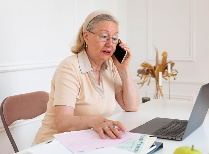 An elderly woman on a phone call, seated at a desk with a laptop and papers, discussing a company decision. An elderly woman on a phone call, seated at a desk with a laptop and papers, discussing a company decision.