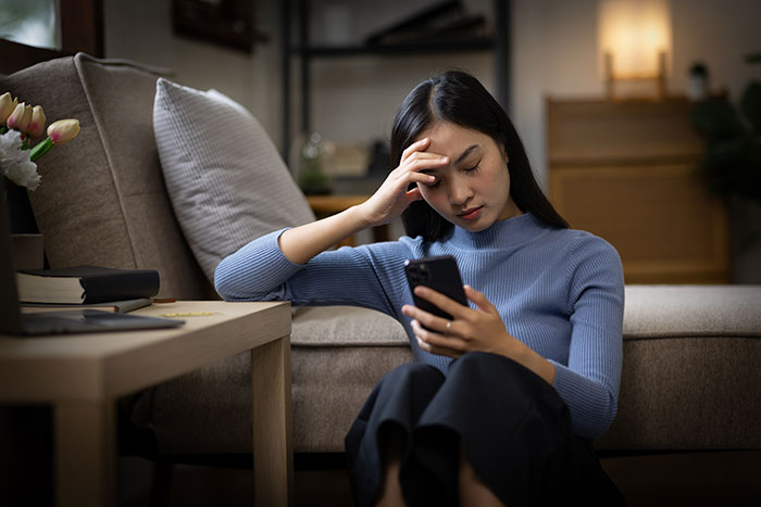 A concerned woman in a blue sweater, seated on the floor and looking at her phone in an apartment. A concerned woman in a blue sweater, seated on the floor and looking at her phone in an apartment.
