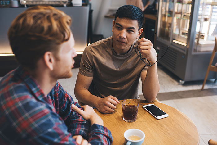 Two men at a café table, one holding glasses and listening, illustrating communication and cultural judgment. Two men at a café table, one holding glasses and listening, illustrating communication and cultural judgment.