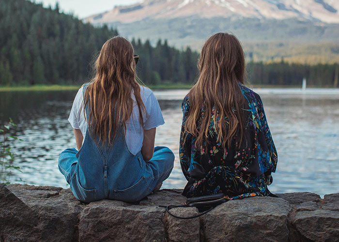 Two women seated by a lake, facing away, possibly discussing a secret that impacts their living situation as roommates. Two women seated by a lake, facing away, possibly discussing a secret that impacts their living situation as roommates.