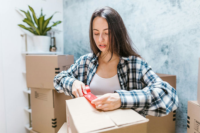 Woman sealing a cardboard box with red tape, standing in a room with several moving boxes and a plant. Woman sealing a cardboard box with red tape, standing in a room with several moving boxes and a plant.