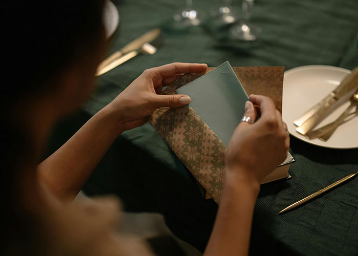 Person opening a wrapped gift at a dining table with green tablecloth and cutlery nearby. Person opening a wrapped gift at a dining table with green tablecloth and cutlery nearby.