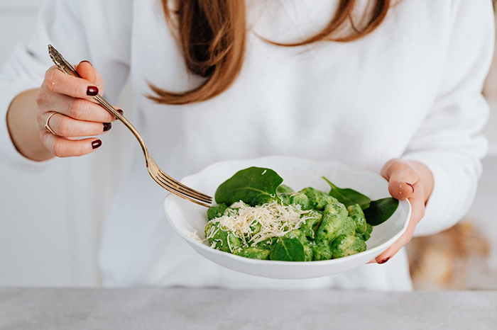 Woman in a white sweater holding a plate of green gnocchi with a fork, garnished with cheese and spinach. Woman in a white sweater holding a plate of green gnocchi with a fork, garnished with cheese and spinach.