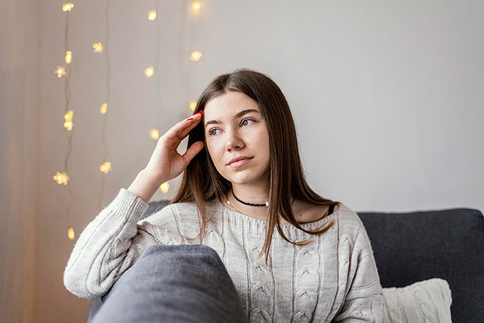 Woman sitting on a gray couch, reflecting, wearing a cream sweater with fairy lights in the background. Woman sitting on a gray couch, reflecting, wearing a cream sweater with fairy lights in the background.