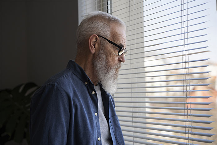 Elderly man in glasses and blue shirt, looking out the window with blinds in a pensive moment. Elderly man in glasses and blue shirt, looking out the window with blinds in a pensive moment.