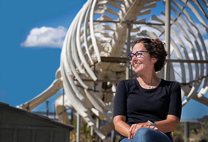 Colossal Biosciences Chief Science Officer Beth Shapiro smiling in front of large dinosaur skeleton under a clear blue sky. Colossal Biosciences Chief Science Officer Beth Shapiro smiling in front of large dinosaur skeleton under a clear blue sky.