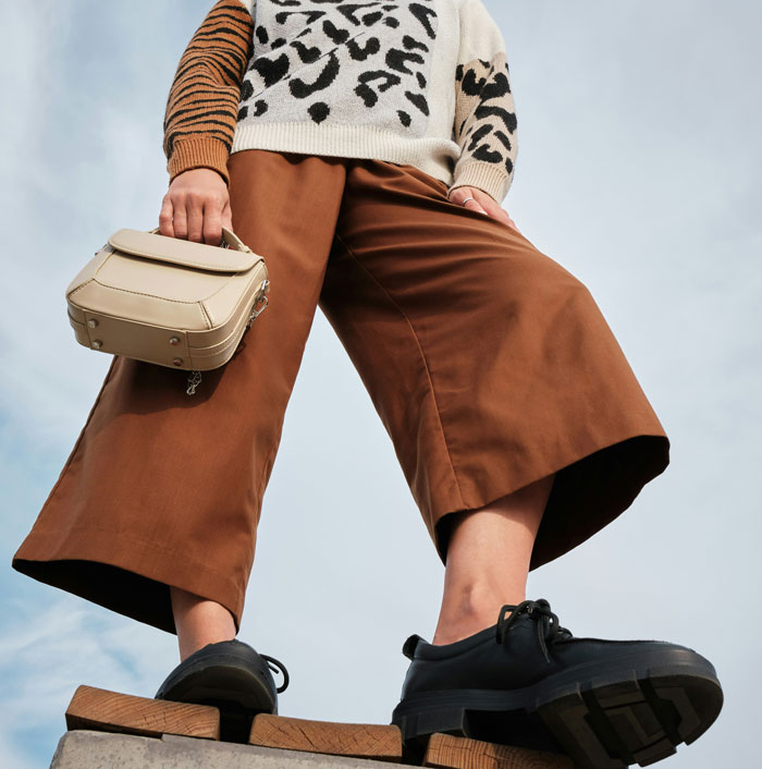 Low-angle view of a person in wide-leg pants, chunky shoes, and leopard print sweater, holding a beige handbag; fashion trends roast.