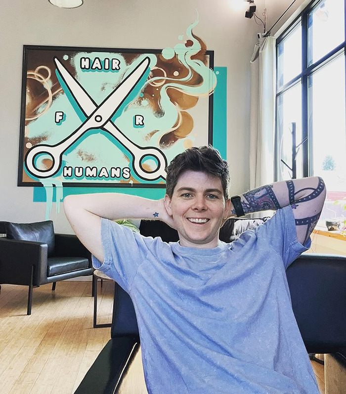 Jules Heron smiling in salon with sign "Hair for Humans," creating an inclusive, safe space for haircuts. Jules Heron smiling in salon with sign "Hair for Humans," creating an inclusive, safe space for haircuts.