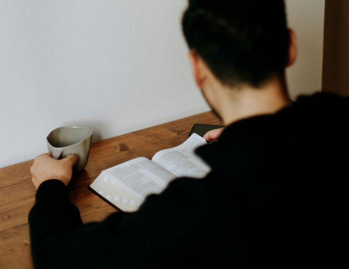 Man reading a book at a wooden table, holding a coffee mug, focusing on finals, not babysitting sister's kids during vacation. Man reading a book at a wooden table, holding a coffee mug, focusing on finals, not babysitting sister's kids during vacation.