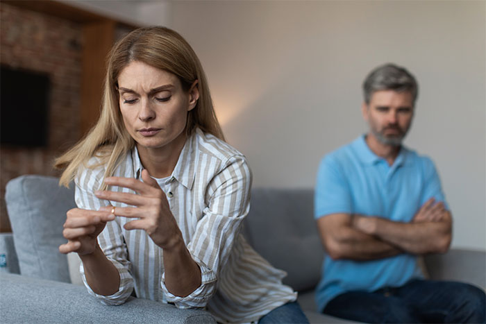 A woman looks at her ring, deep in thought, while a man in a blue shirt sits in the background, arms crossed, highlighting ex-husband regrets. A woman looks at her ring, deep in thought, while a man in a blue shirt sits in the background, arms crossed, highlighting ex-husband regrets.