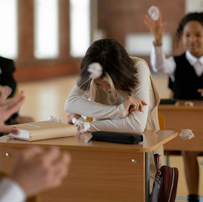 Teen looking uncomfortable in a classroom setting, covering head while being targeted with paper balls. Teen looking uncomfortable in a classroom setting, covering head while being targeted with paper balls.