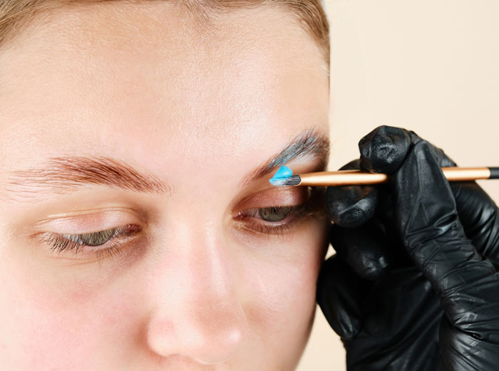 Person applying blue color to eyebrow with brush, highlighting fashion trends critique.