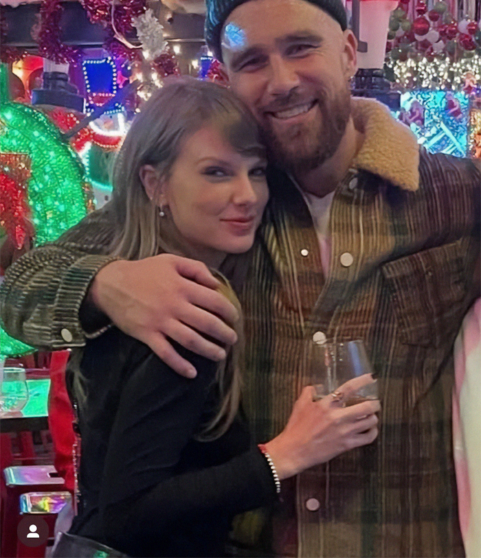 Taylor Swift smiling at an event, wearing a black outfit, amid festive decor, with Travis Kelce in a plaid jacket. Taylor Swift smiling at an event, wearing a black outfit, amid festive decor, with Travis Kelce in a plaid jacket.