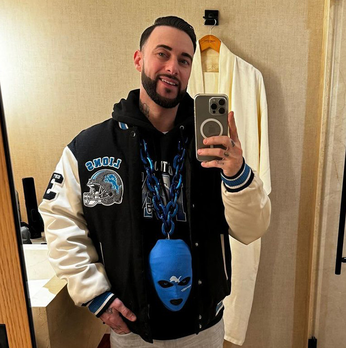 Man taking a mirror selfie in a bathroom, wearing a Lions jacket and holding a phone with a prominent blue tag accessory. Man taking a mirror selfie in a bathroom, wearing a Lions jacket and holding a phone with a prominent blue tag accessory.