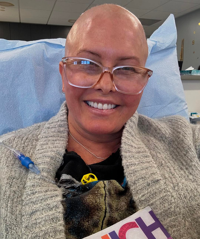 Nicole Eggert in hospital setting smiling, wearing glasses and cozy sweater, highlighting cancer journey. Nicole Eggert in hospital setting smiling, wearing glasses and cozy sweater, highlighting cancer journey.
