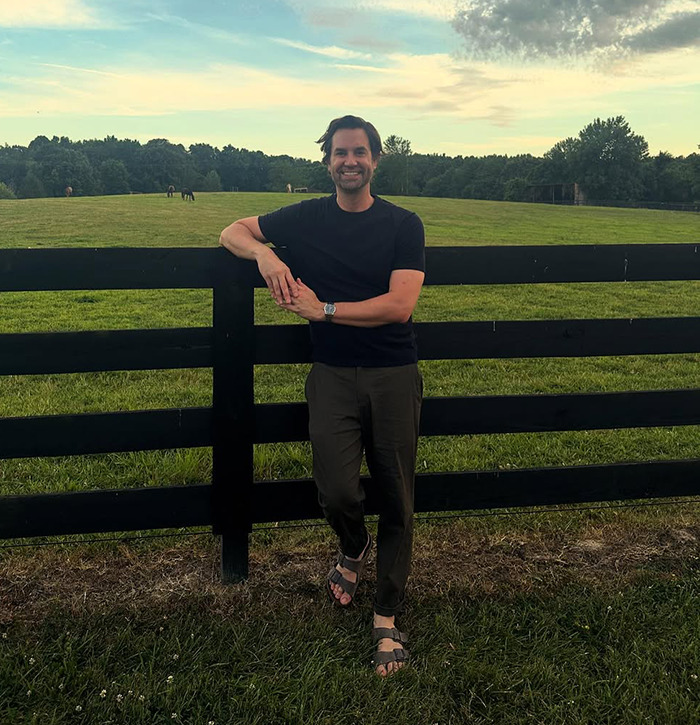 Man smiling and leaning on a black fence in a green field. Man smiling and leaning on a black fence in a green field.