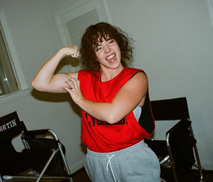 Sydney Sweeney in a red tank top flexing her bicep, smiling at the camera in a casual indoor setting. Sydney Sweeney in a red tank top flexing her bicep, smiling at the camera in a casual indoor setting.