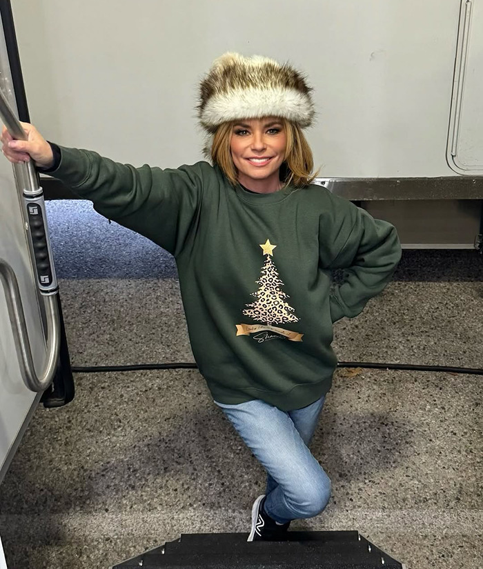 Shania Twain in festive attire posing on steps, wearing a green sweater with a printed Christmas tree and a fur hat. Shania Twain in festive attire posing on steps, wearing a green sweater with a printed Christmas tree and a fur hat.
