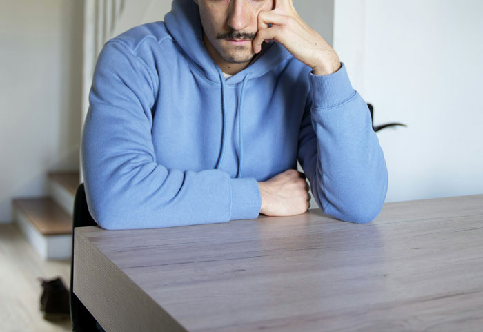 Man in blue hoodie, looking reflective at a wooden table, relating to a family conflict involving a stepdad. Man in blue hoodie, looking reflective at a wooden table, relating to a family conflict involving a stepdad.