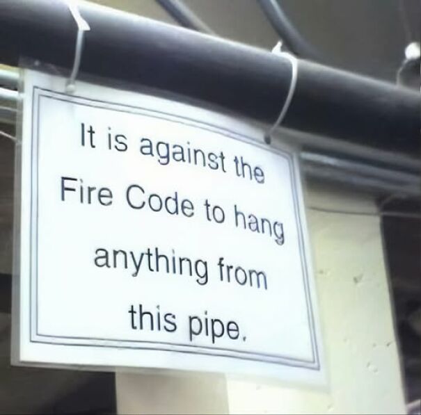 Weird and absurd sign stating it's against the Fire Code to hang items from a pipe in a building.