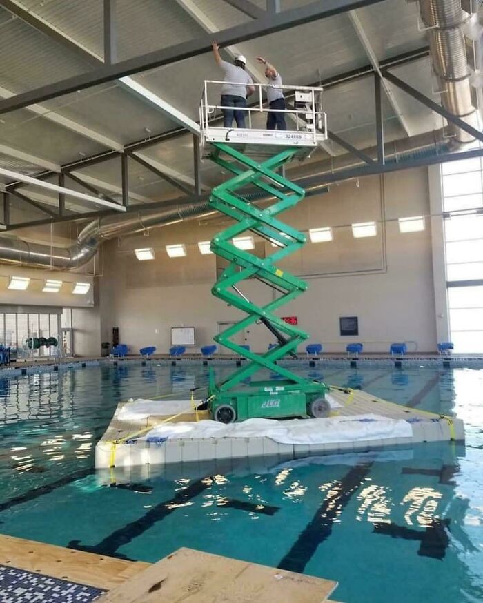 Two employees blatantly ignoring safety protocol by standing on a scissor lift platform floating on a pool.